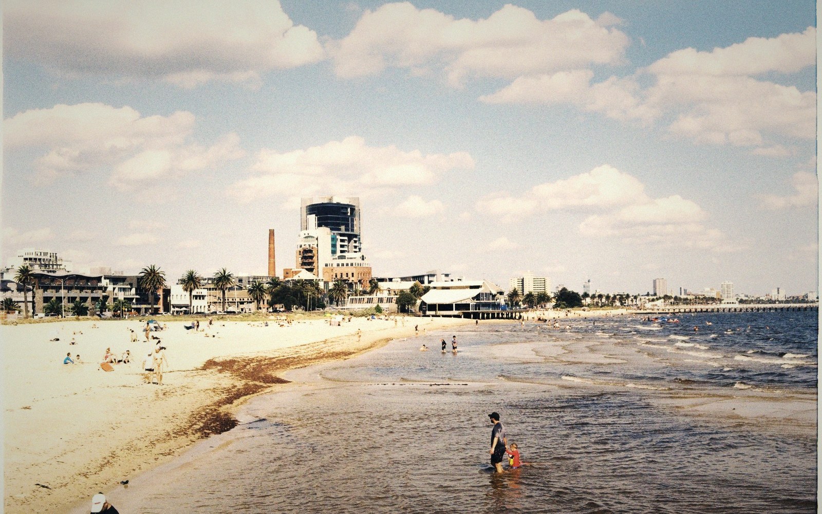 People wade in shallow water along a sandy beach with a city skyline behind them