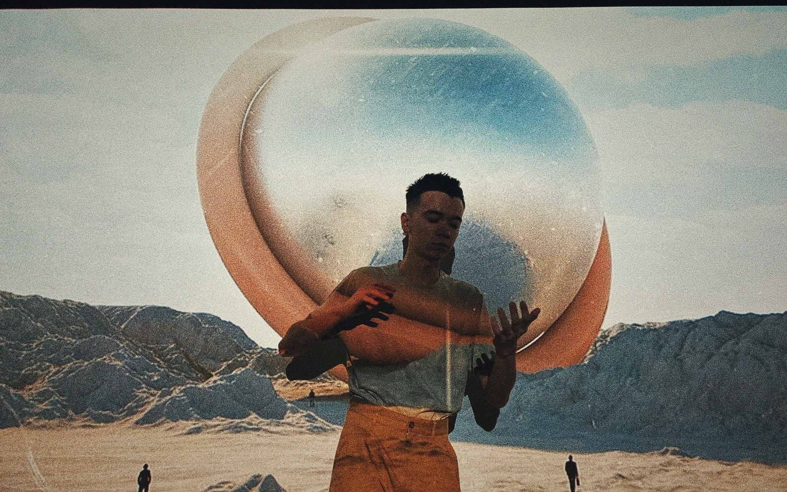 A man stands before a large sphere in a desert landscape, gesturing with open hands