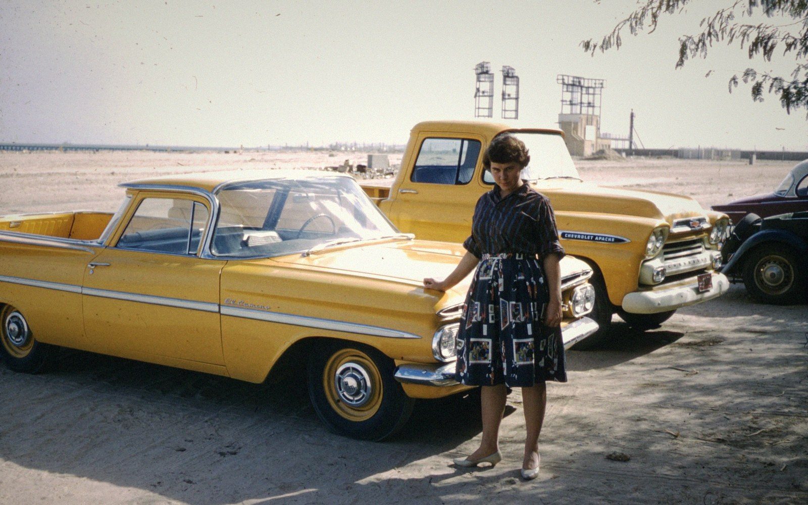 Woman in a patterned dress stands beside a yellow El Camino and Chevrolet Apache truck