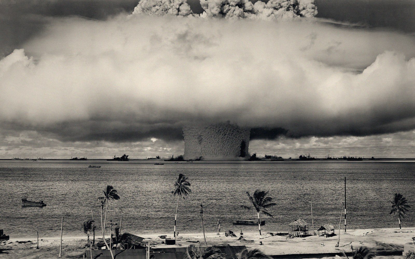 A massive mushroom cloud rises above the ocean beyond a palm-lined shore