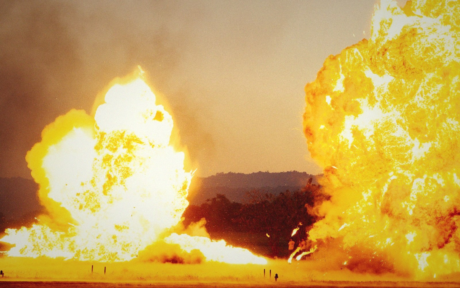 Two massive fireballs erupt from the ground with silhouetted hills in the background